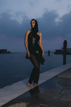 Model standing on a stone pier by the water at dusk wearing a black sequined dress and head covering