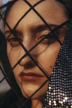 Close-up portrait of a woman's face partially obscured by a dark mesh veil with a diamond pattern.