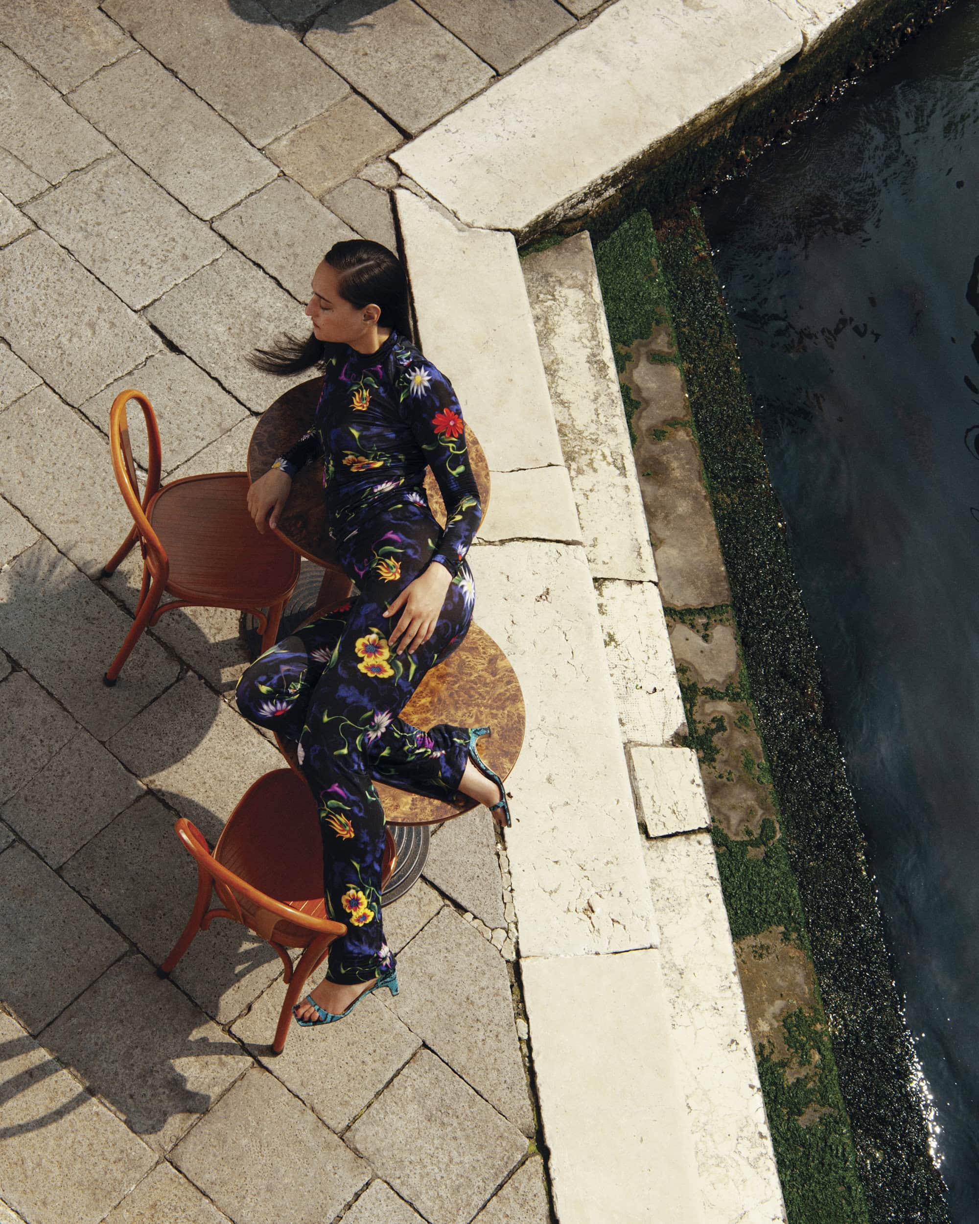 High-angle fashion photograph of a model in a floral patterned jumpsuit seated on a round café table by a stone canal edge.
