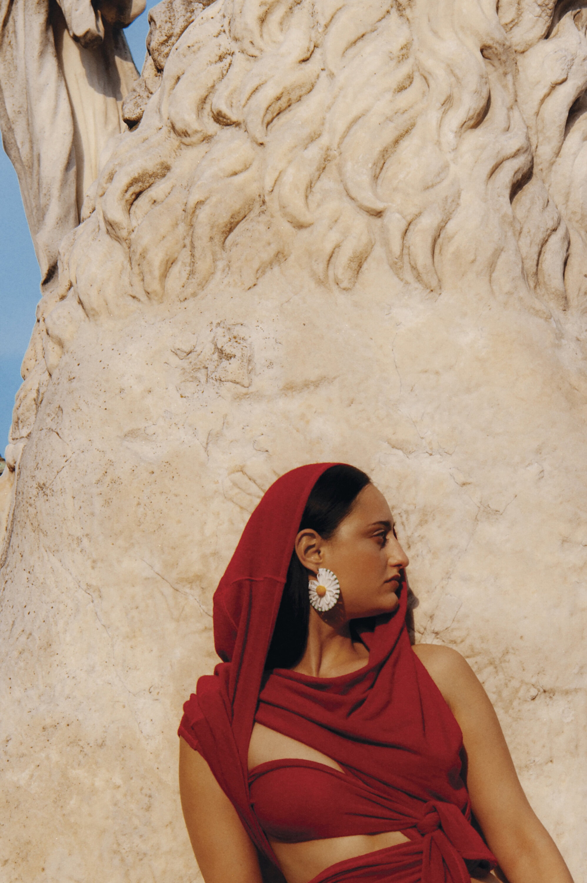 Woman wearing red hooded top and floral earring posing against weathered stone sculpture in Venice