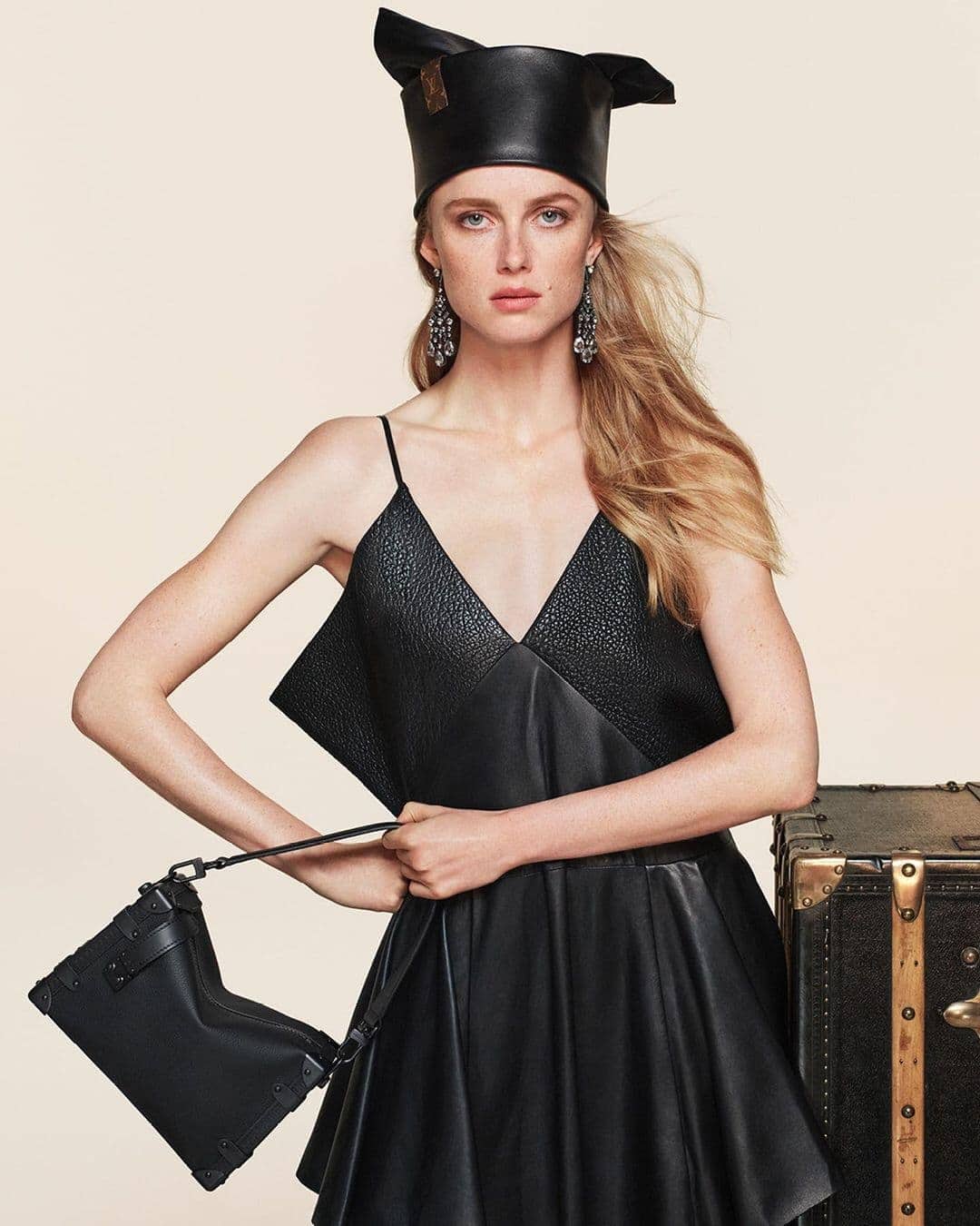 Model Rianne Van Rompaey in a black leather dress holding a handbag against a plain background