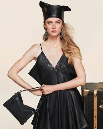 Model Rianne Van Rompaey in a black leather dress holding a handbag against a plain background