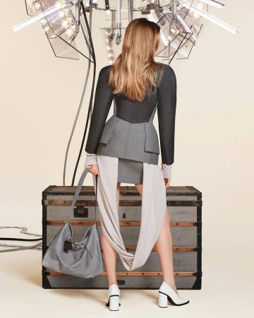 Model seen from behind wearing structured grey jacket and skirt holding a leather shoulder bag in front of a vintage trunk