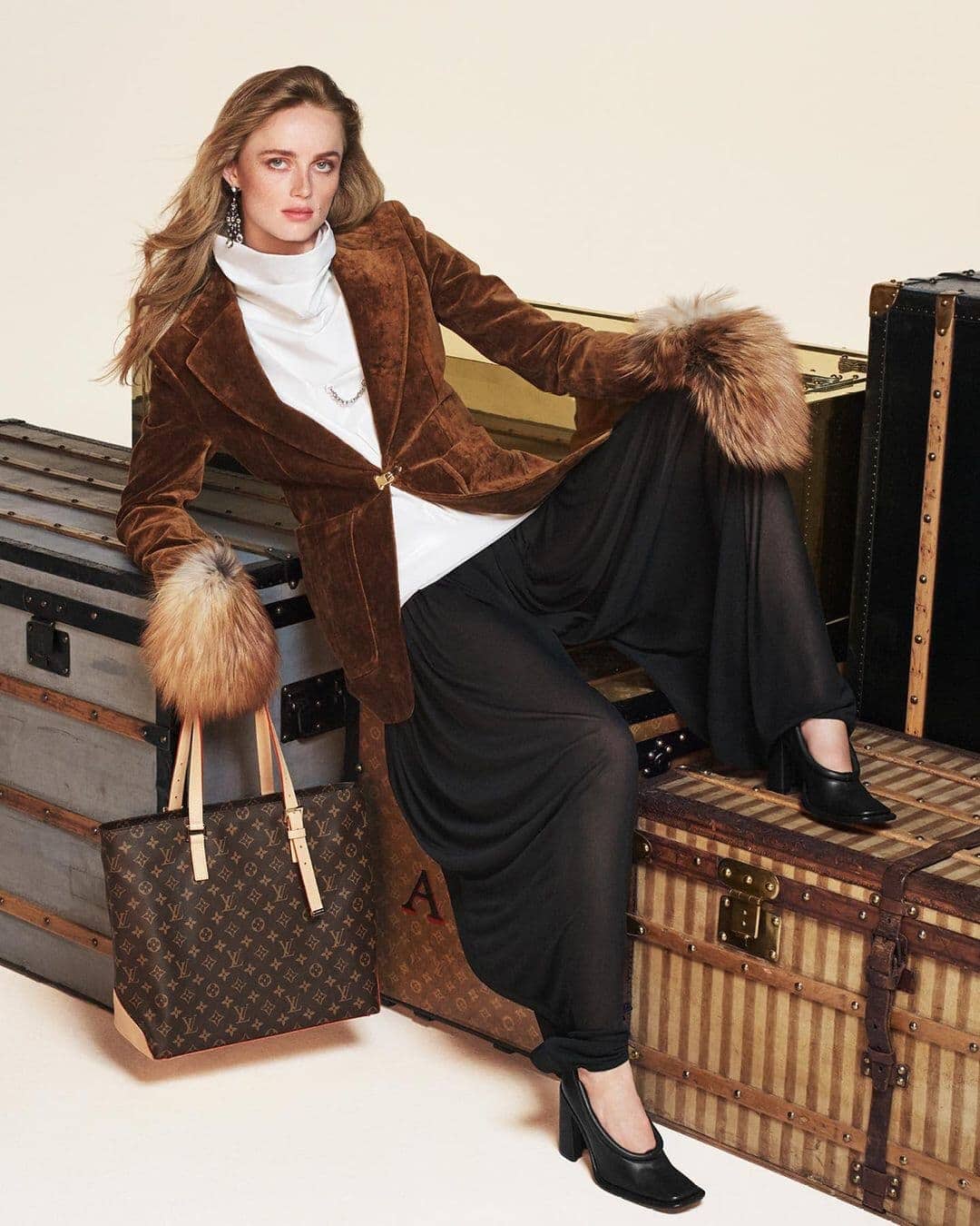 Model wearing brown jacket with fur cuffs and black trousers sitting on a vintage trunk next to a monogram tote bag