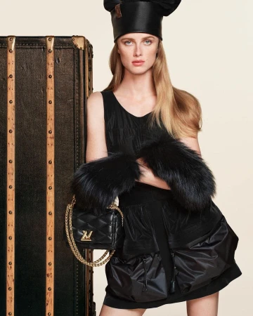 Model wearing black dress and matching hat leaning against a large dark trunk for Louis Vuitton Fall 2024 ad campaign