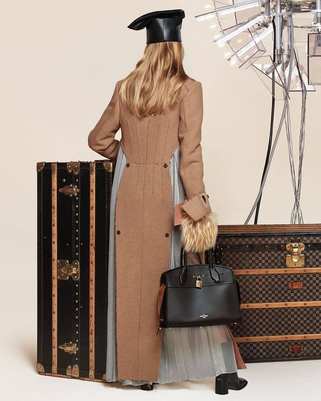 Model standing facing away, wearing a structured camel coat and black leather hat, holding a black handbag near vintage trunks