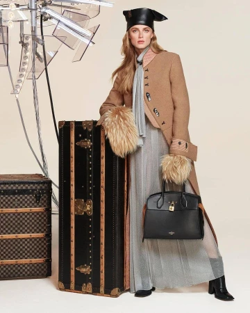 Model wearing a tan coat and black hat leaning on a large vintage Louis Vuitton trunk with a black handbag.
