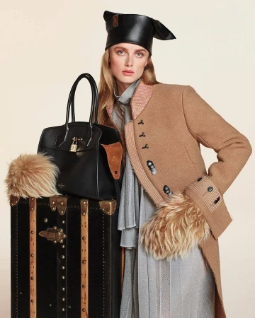 Rianne Van Rompaey wearing a camel coat and black leather hat leaning on a vintage trunk with a black handbag for Louis Vuitton Fall 2024.