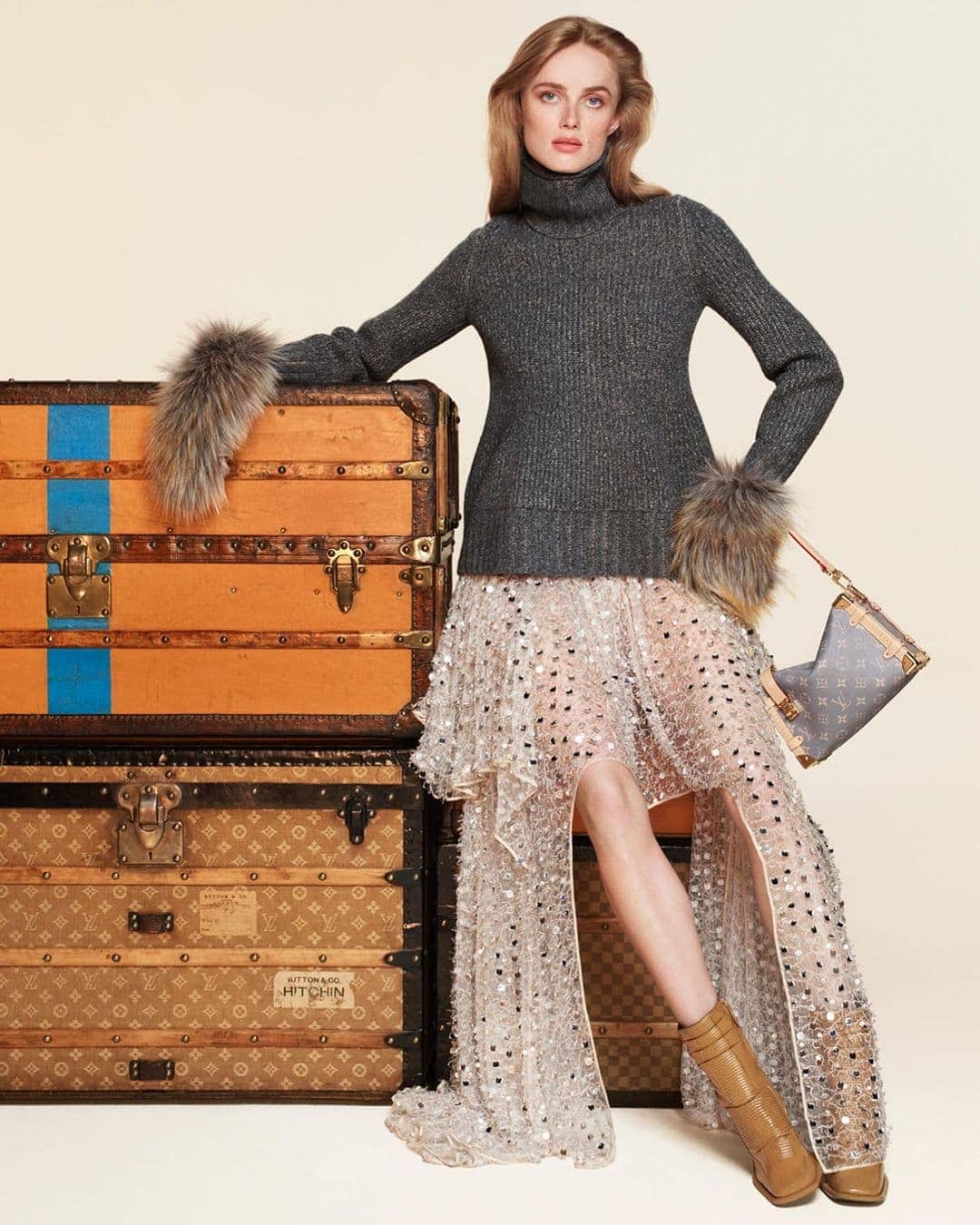 Model leaning on stacked vintage trunks wearing a dark turtleneck and sequined skirt for Louis Vuitton