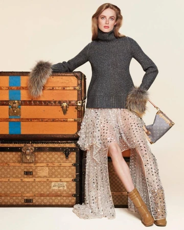 Model leaning on stacked vintage trunks wearing a dark turtleneck and sequined skirt for Louis Vuitton