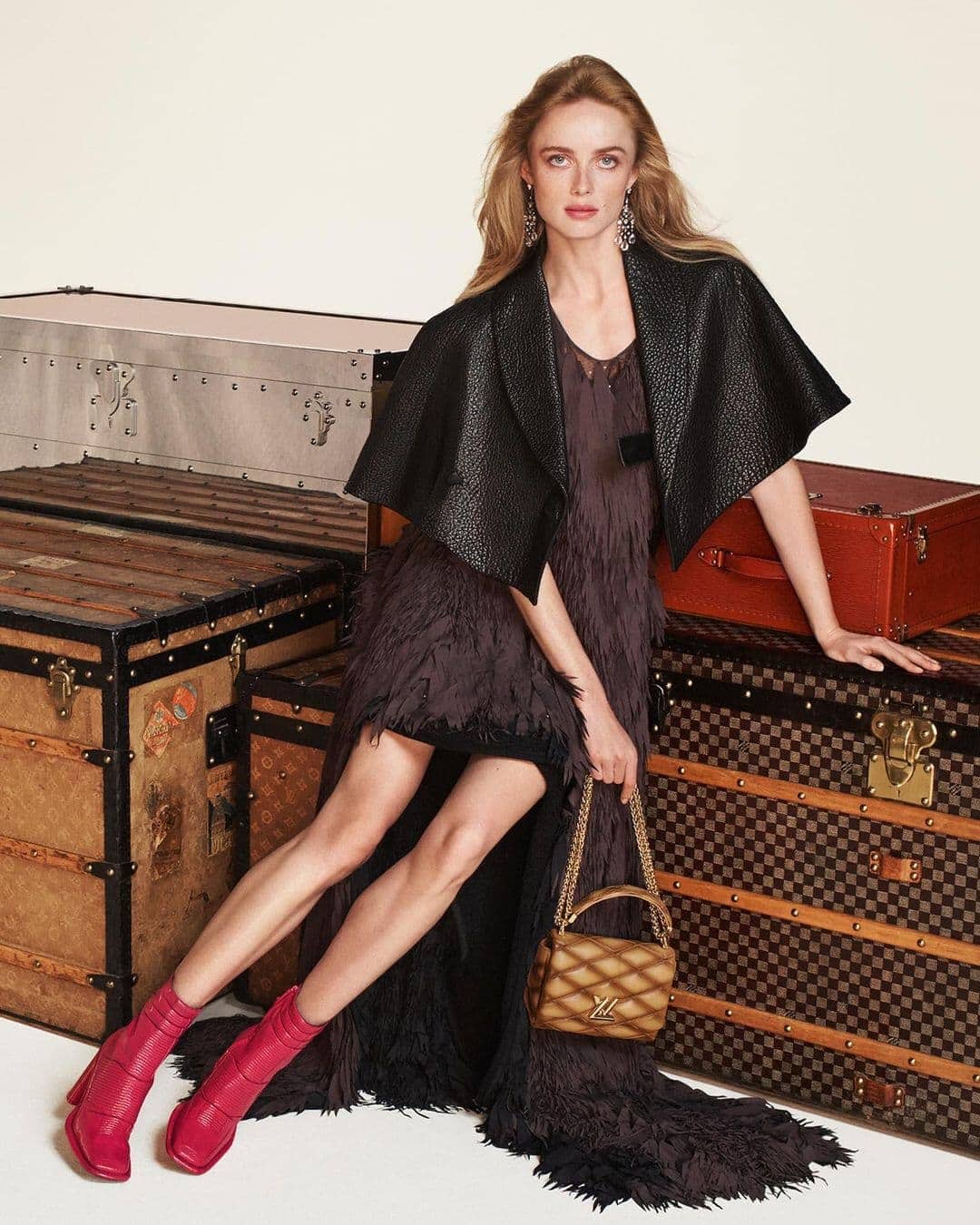 Model Rianne Van Rompaey seated amongst vintage Louis Vuitton trunks wearing a dark cape, fringe dress, and pink boots.