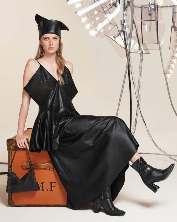 Model in black leather dress and hat sitting on a vintage trunk holding a handbag against a plain background
