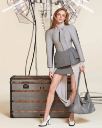 Model wearing grey structured jacket and mini skirt holding a grey handbag standing in front of a vintage trunk.