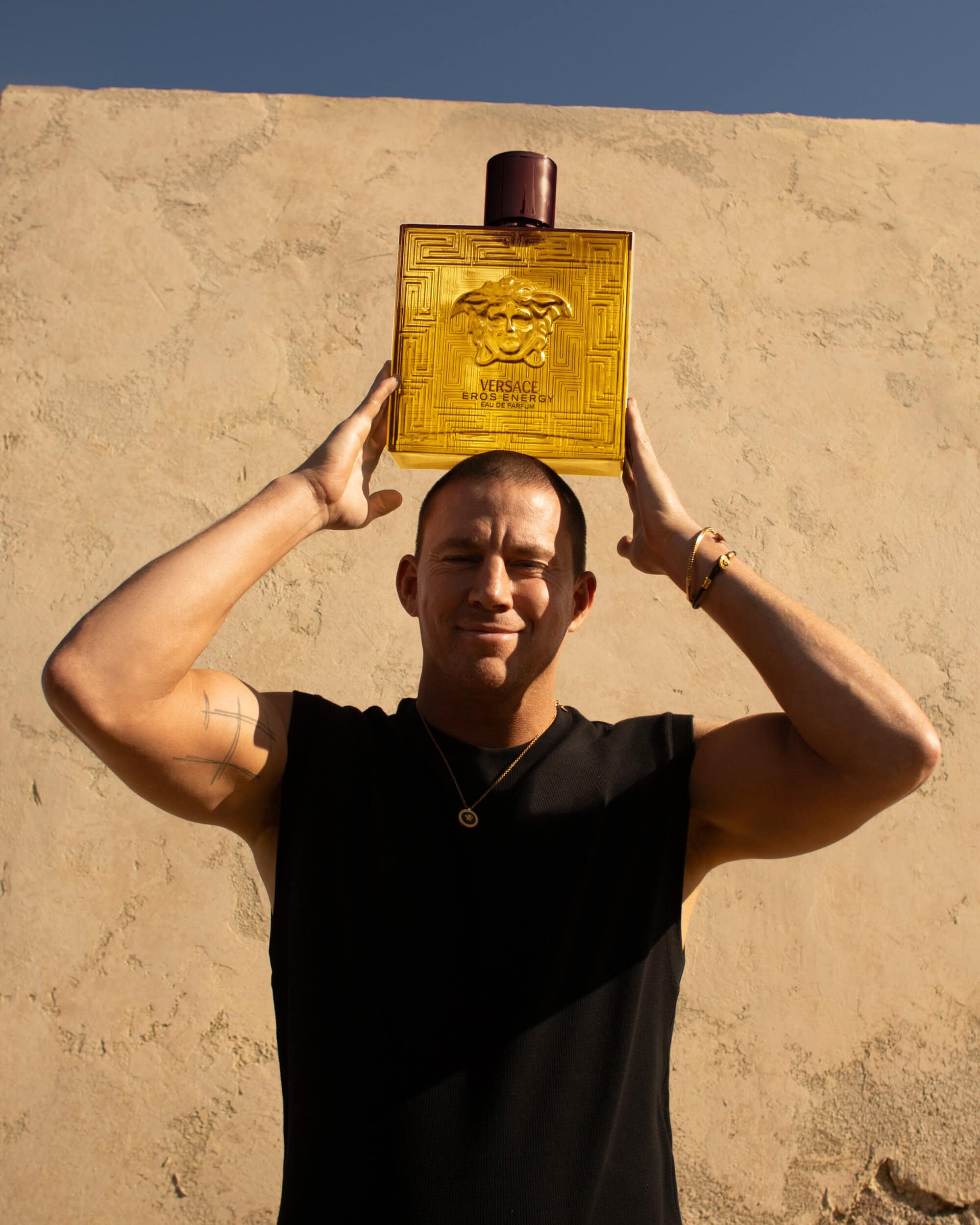 Channing Tatum holding a large Versace Eros Energy perfume bottle above his head against a stucco wall
