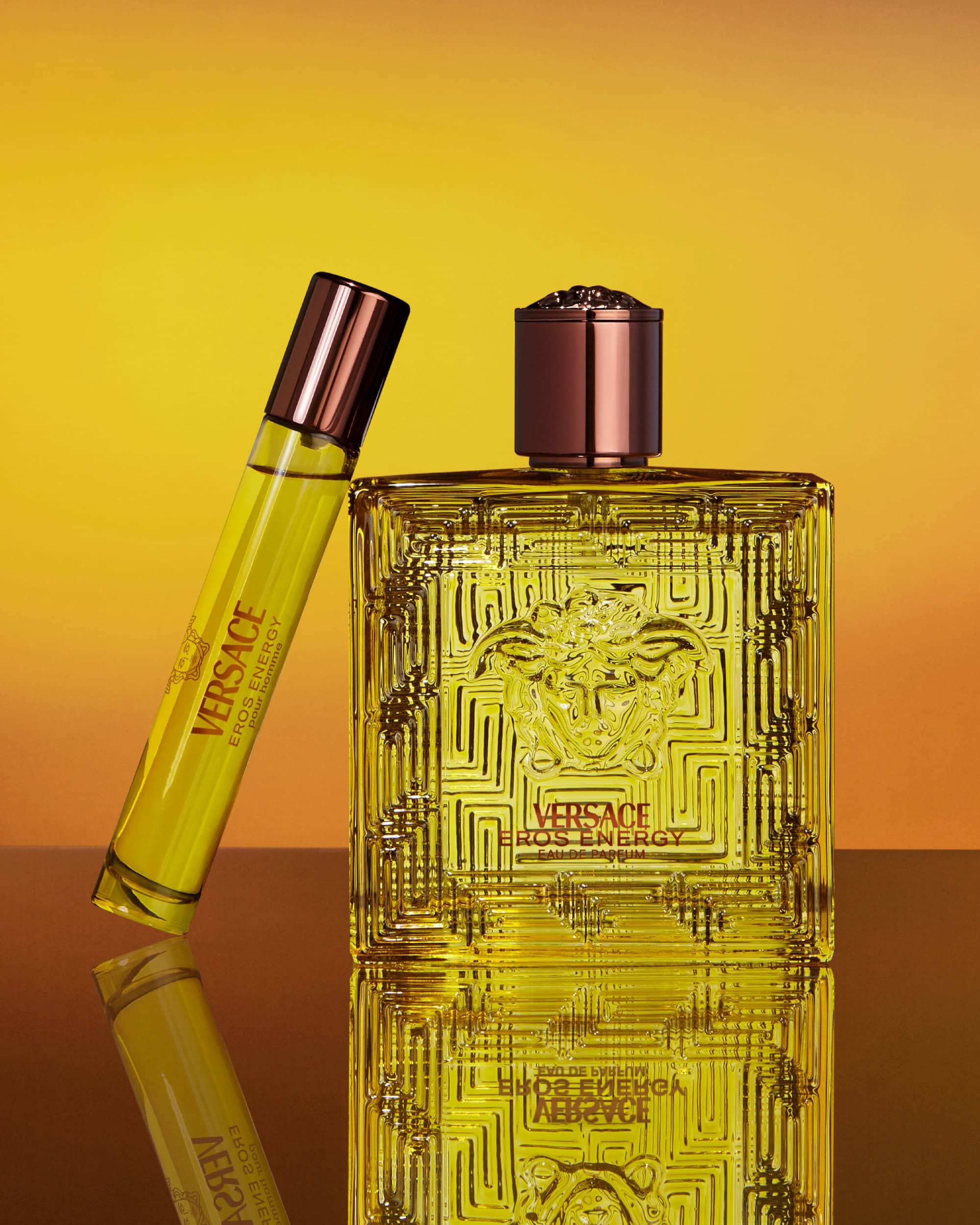 Versace Eros Energy fragrance bottle and travel spray standing on a reflective surface against a yellow gradient background