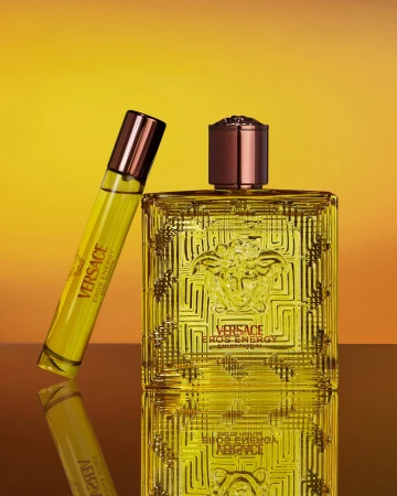 Versace Eros Energy fragrance bottle and travel spray standing on a reflective surface against a yellow gradient background