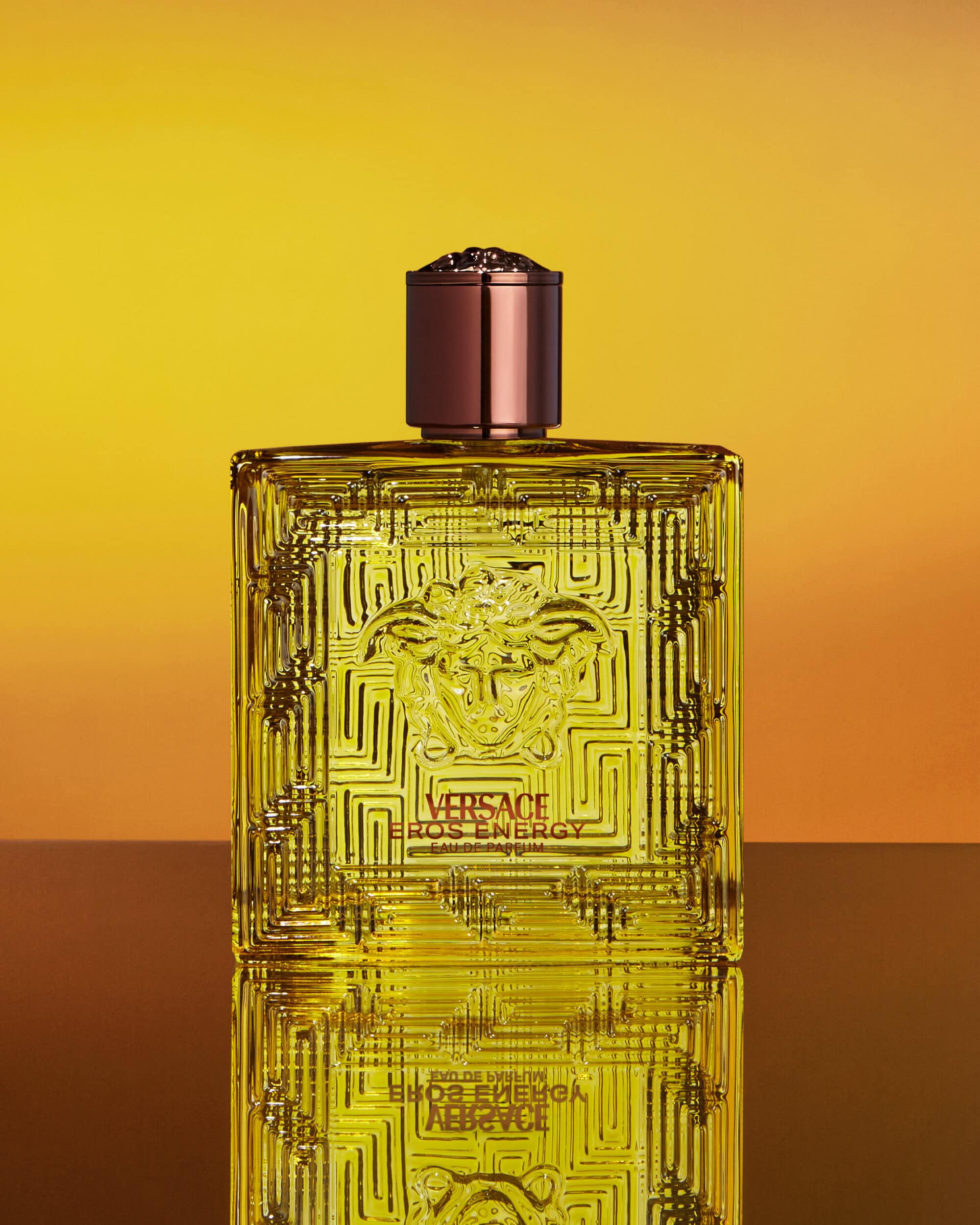 Versace Eros Energy perfume bottle centered against a warm gradient yellow background with a reflective surface below