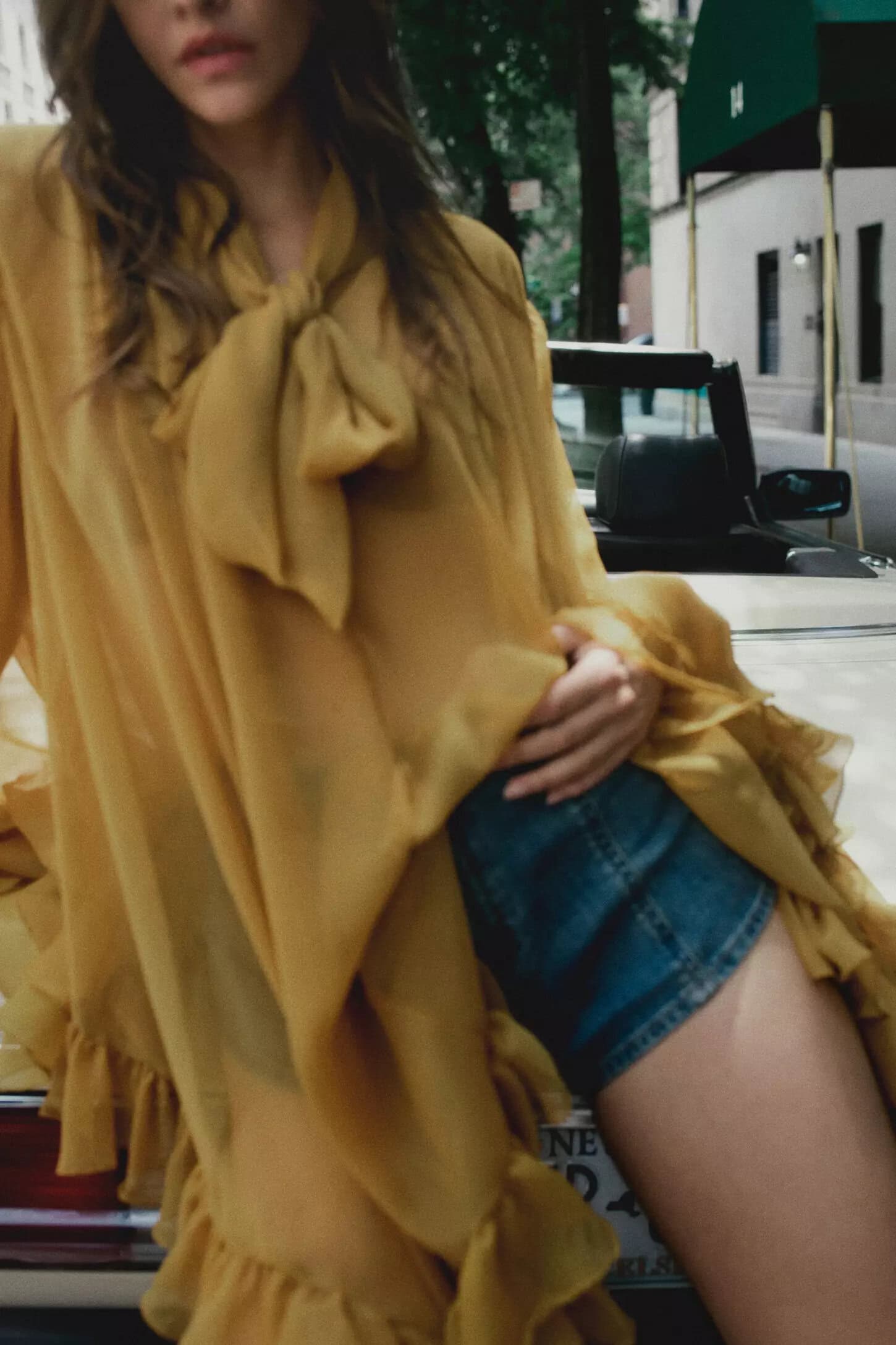 Model in a yellow blouse with bow tie and denim shorts leaning against a vintage car on a city street