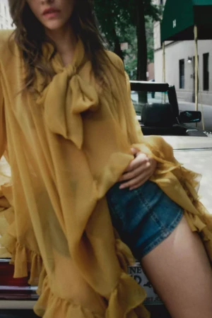 Model in a yellow blouse with bow tie and denim shorts leaning against a vintage car on a city street