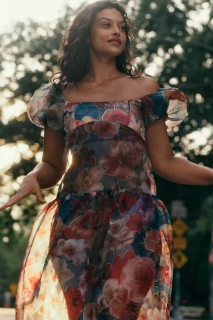 Model wearing a sheer floral print off-the-shoulder dress outdoors among trees during the Fall 2024 campaign for Anthropologie.