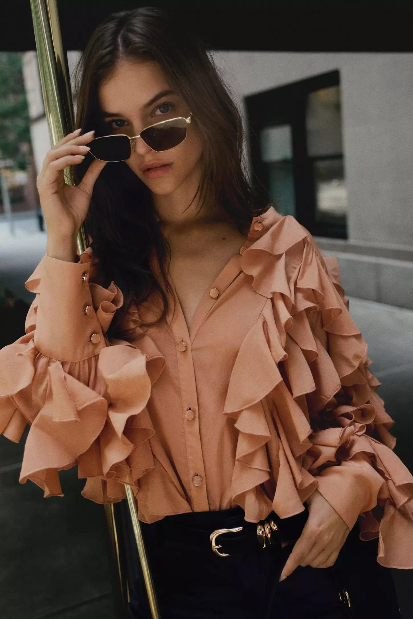 Barbara Palvin wearing a peach ruffled blouse and sunglasses leaning against a pole outdoors for Anthropologie.