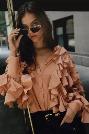 Barbara Palvin wearing a peach ruffled blouse and sunglasses leaning against a pole outdoors for Anthropologie.