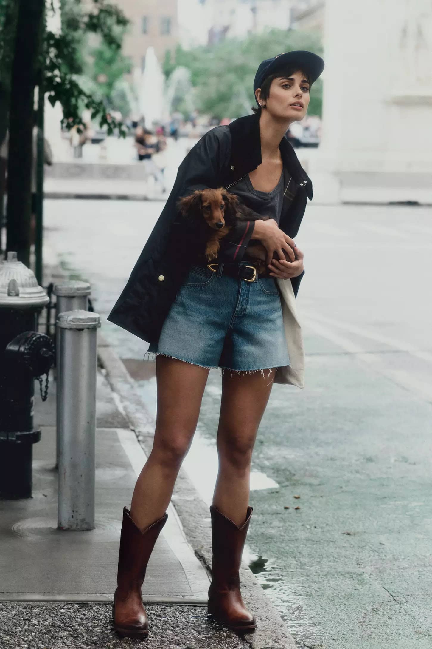 Model standing on a city street holding a dachshund dog while wearing denim shorts, cowboy boots, and a jacket.