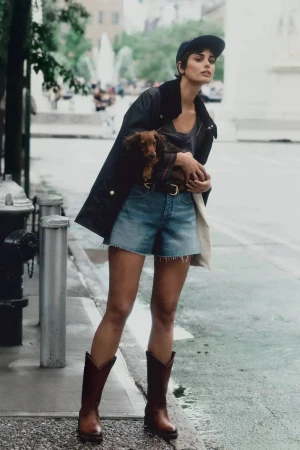 Model standing on a city street holding a dachshund dog while wearing denim shorts, cowboy boots, and a jacket.
