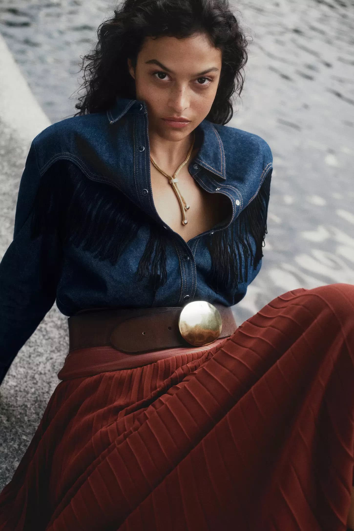Model wearing a denim fringed shirt and pleated red skirt outdoors near water for Anthropologie Fall 2024 campaign