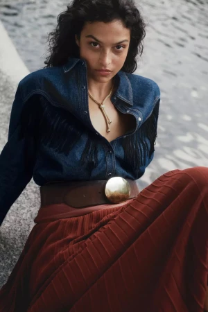 Model wearing a denim fringed shirt and pleated red skirt outdoors near water for Anthropologie Fall 2024 campaign
