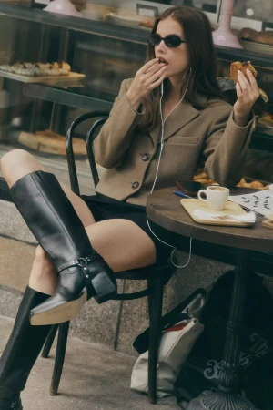 Model wearing a brown blazer and black boots eating food at an outdoor café table for Anthropologie Fall 2024 campaign