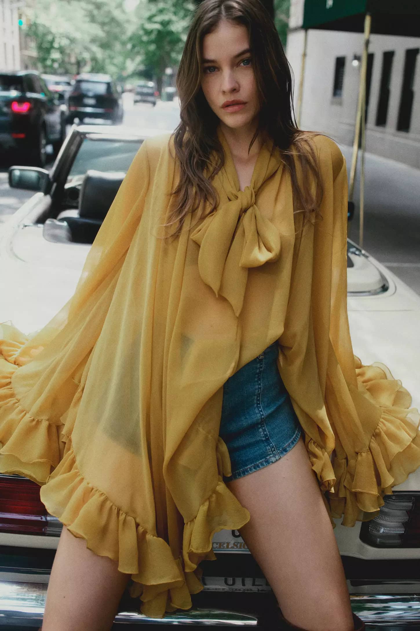 Barbara Palvin leaning on a white convertible car in a yellow blouse and denim shorts on a New York City street.