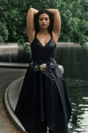 Model in a black dress with a studded heart belt posing outdoors by water