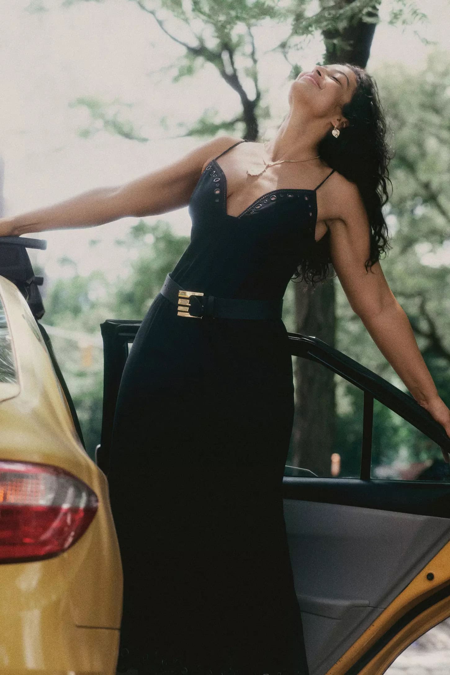 Woman in a black dress leaning against a yellow taxi in an outdoor setting