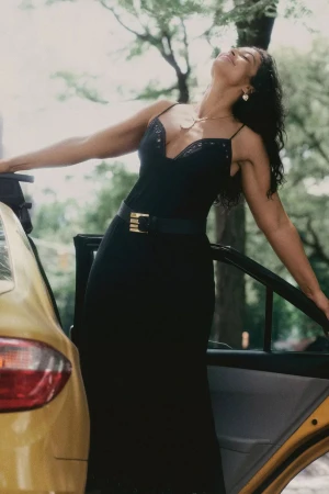Woman in a black dress leaning against a yellow taxi in an outdoor setting
