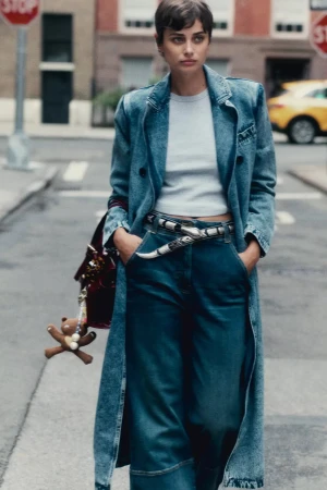Model in denim coat and jeans standing on a city street holding a handbag