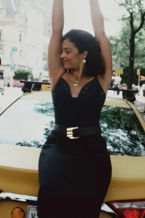 Woman in a black dress leaning against a yellow taxi cab on a city street