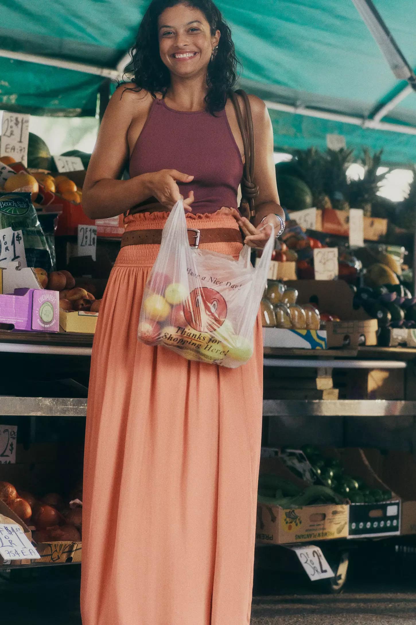 Smiling woman holding a plastic bag of fruit at a market stall, wearing a sleeveless purple top and long peach skirt.