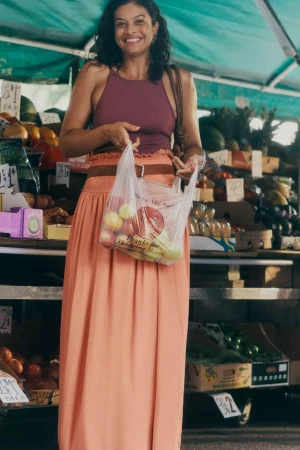 Smiling woman holding a plastic bag of fruit at a market stall, wearing a sleeveless purple top and long peach skirt.
