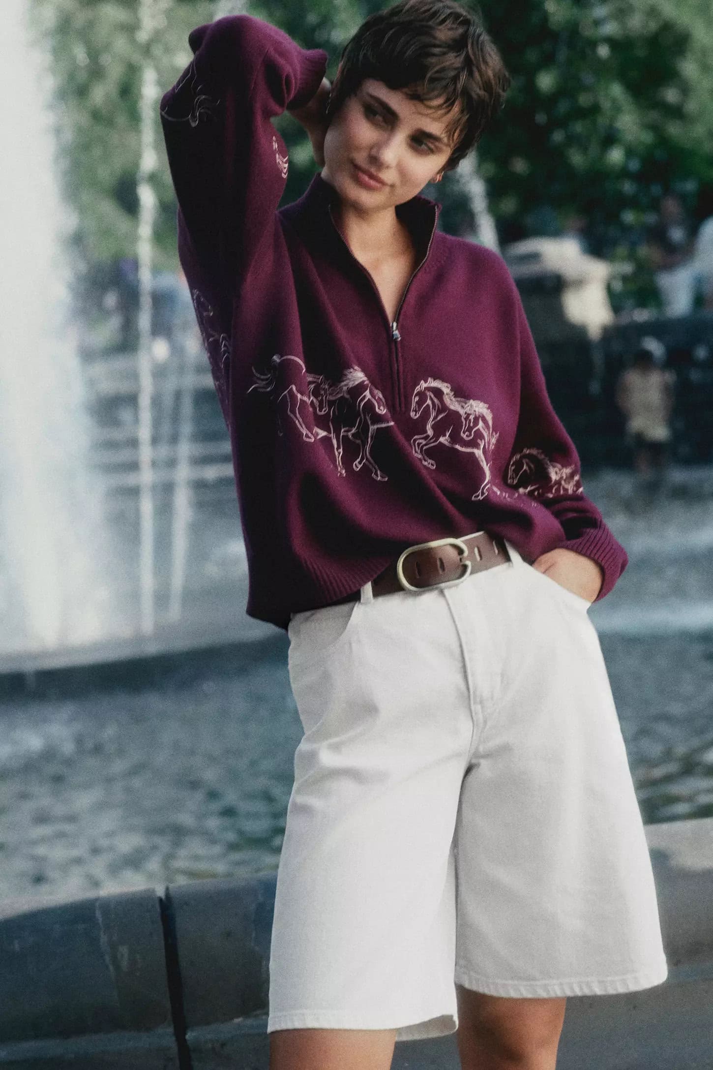 Woman with short dark hair wearing a plum zip-up sweater and white shorts posing outdoors near a fountain.