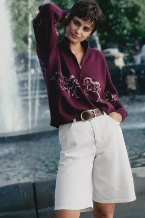 Woman with short dark hair wearing a plum zip-up sweater and white shorts posing outdoors near a fountain.