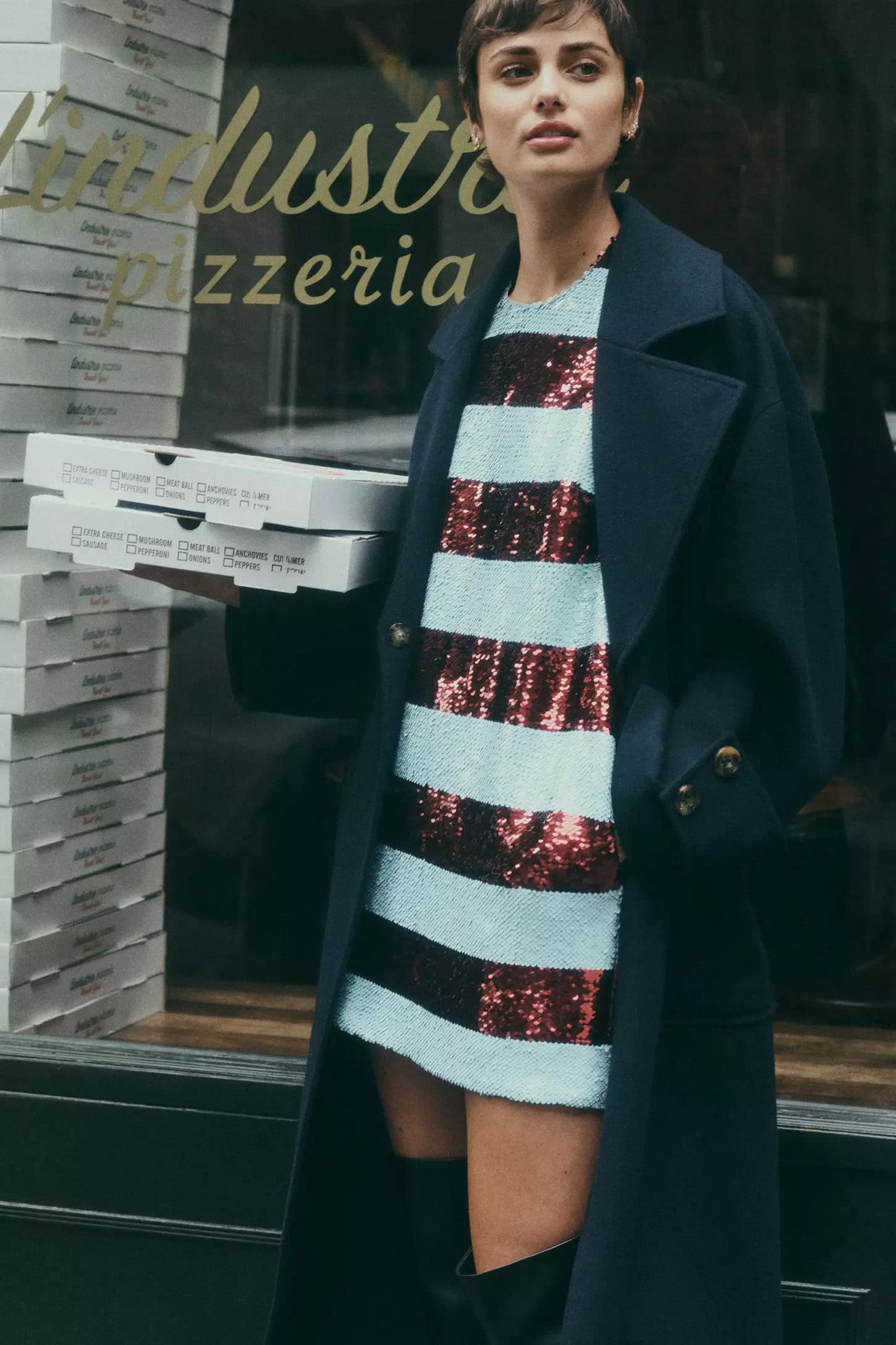 Woman in dark coat and sequin dress holding pizza boxes outside a restaurant