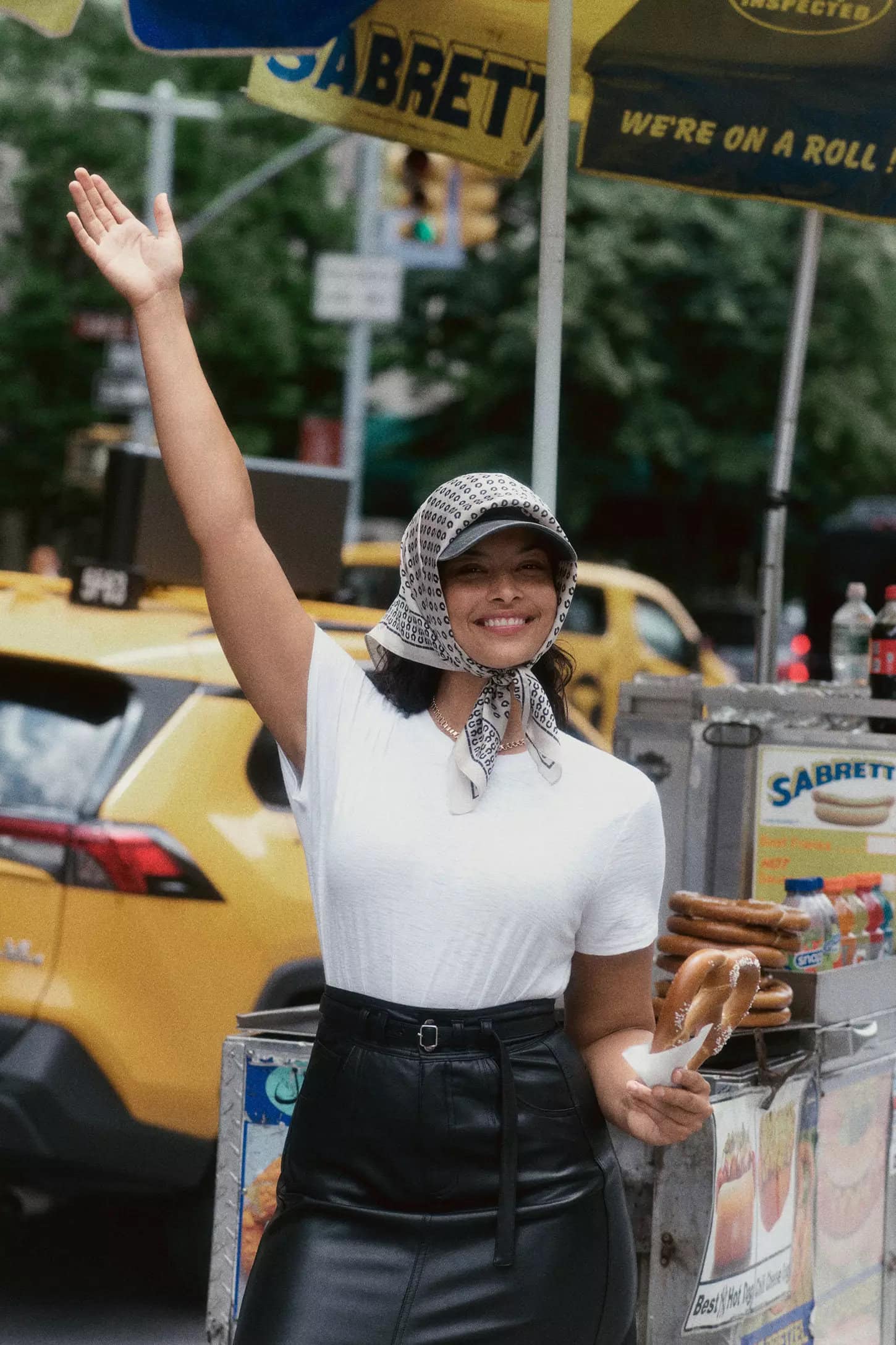 Model waving near a pretzel cart wearing a white t-shirt, black skirt, patterned headscarf, and holding a pretzel in New York City