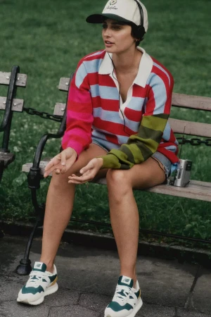 Woman wearing a multi-coloured striped polo sweater and baseball cap seated on a park bench