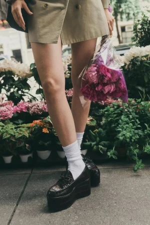 Close-up of legs wearing platform loafers, white socks, and a tan jacket, holding a bouquet of pink flowers outdoors.