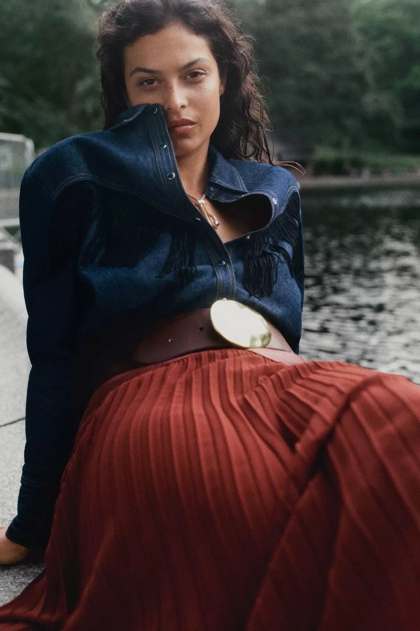 Model wearing a blue denim shirt and rust pleated skirt seated outdoors near water for Anthropologie Fall 2024 campaign