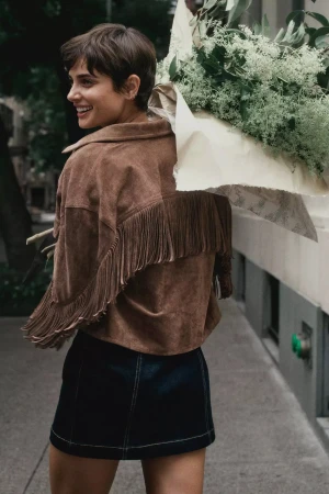 Smiling woman carrying large bouquet of white flowers wearing brown fringed suede jacket and dark skirt outdoors on city sidewalk.