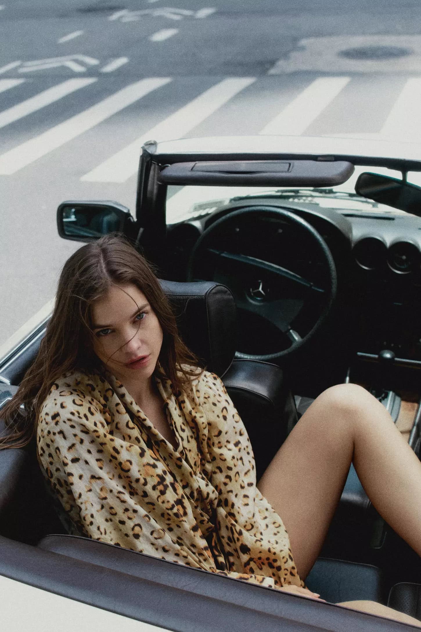Model Barbara Palvin wearing a leopard print shirt seated in a vintage convertible car on a street.