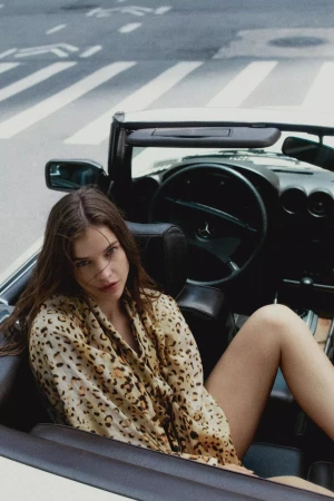 Model Barbara Palvin wearing a leopard print shirt seated in a vintage convertible car on a street.