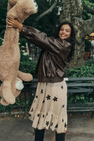 Smiling woman in a brown leather jacket holding up a large stuffed animal outdoors near a park bench